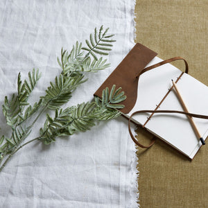 Artificial silver wattle leaf decor arranged with a leather-bound notebook and pencil on a textured surface
