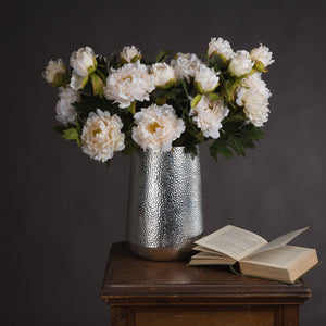 Artificial white peony arrangement in a silver vase on a wooden table next to an open book